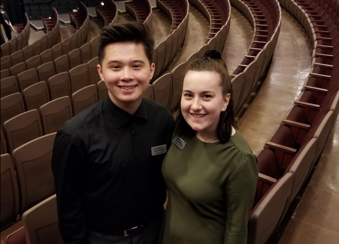 Two smiling student house managers stand in Essex Hall with rows of seating visible behind them