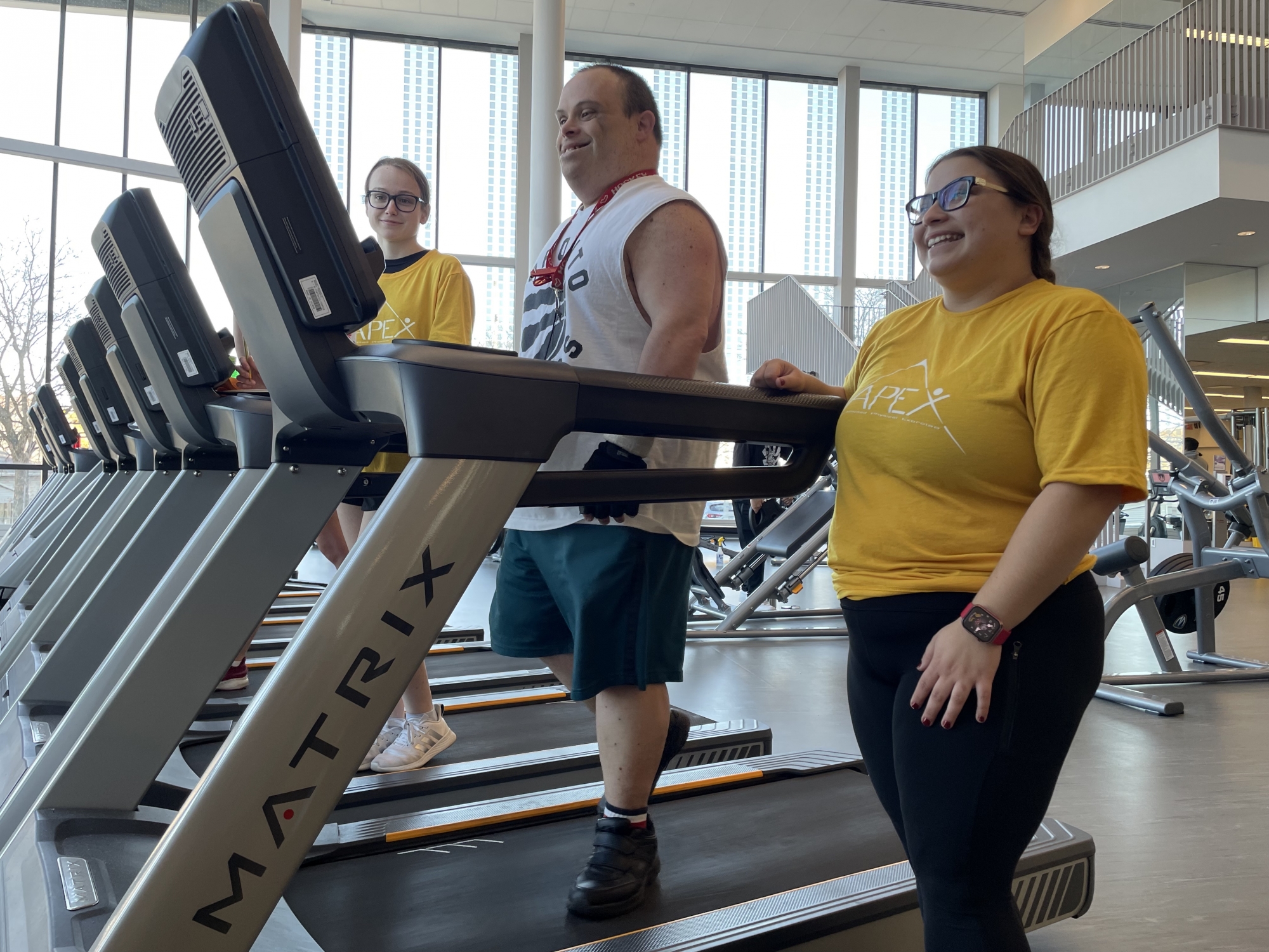 A man walks on a treadmill while two female trainers in yellow T-shirts look on.