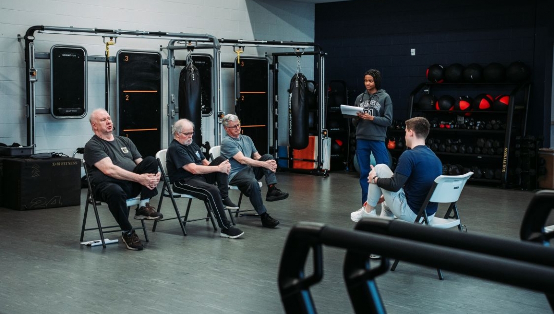 three older men do chair stretches across from a UWindsor student with another student observing with a clipboard