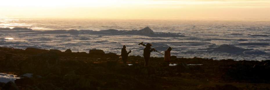 Three unidentified UWindsor researchers trek across a rocky shoreline at sunset, carrying varous measuring equipment.