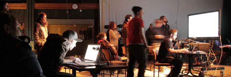 Severel students in a busy, darkened SoCa laboratory/working space pay close attention to illuminated screens and other equipment