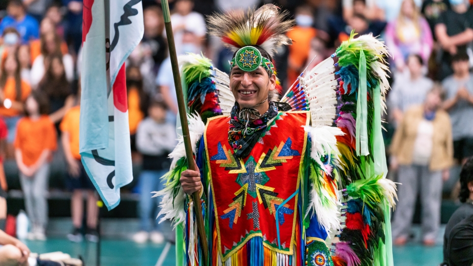 An Indigenous man wearing colourful traditional Pow Wow regalia holds a flag.