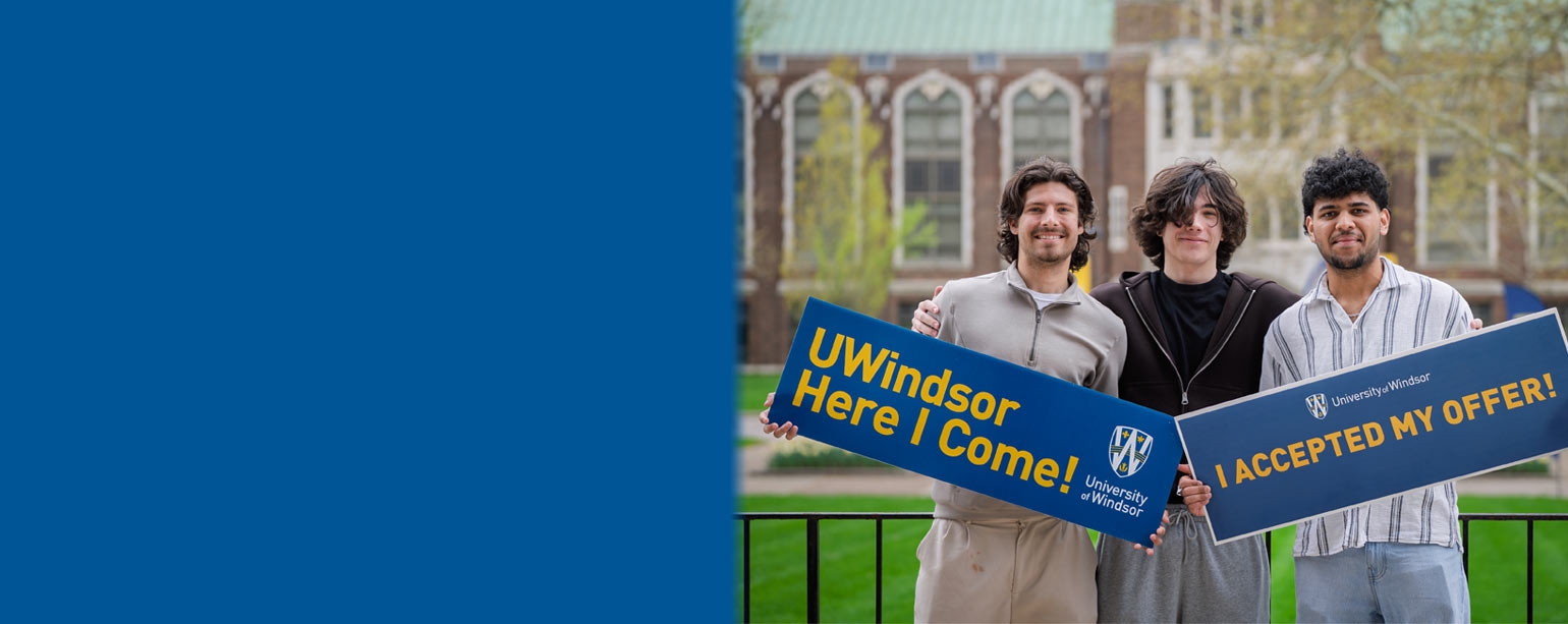Three male students standing in front of Dillon Hall holding signs saying they were accepted to UWindsor.