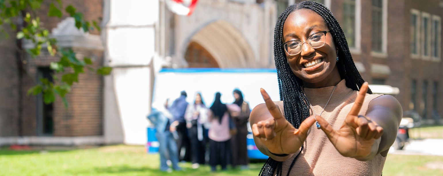 Girl standing by Dillon Hall with her hands and fingers shaped like a W