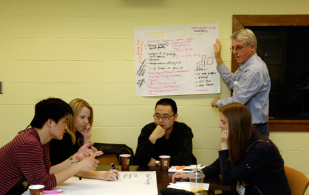 Damian Ruth stands beside a large piece of paper taped to a wall with writing all over it. Four younger people sit at a table in front of him.