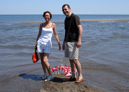 Marianne Poumay and François Georges posing at the southernmost tip of Canada. On the ground beneath them are numerous small Canadian flags planted into the sand.