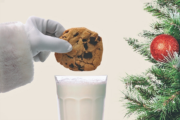 gloved hand dunking cookie in milk next to decorated tree