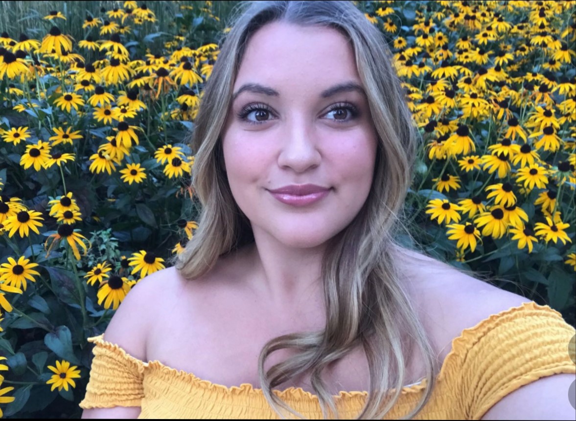 This is an upper body shot of Kat Muscedere. Kat is in a field of yellow flowers and she is smiling in the camera. 