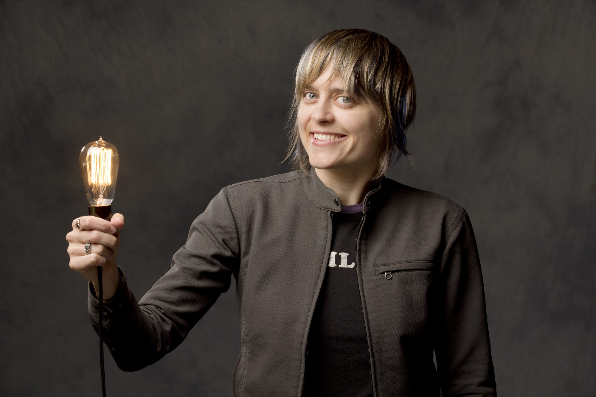 Kirsten Watt, Lighting Designer. Smiling and holding up an illuminated light bulb.