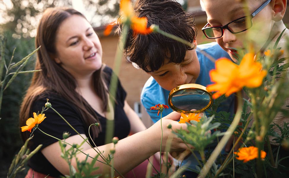 Female student teacher holding a magnifying class at a garden with science students 