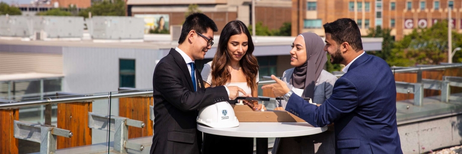 Group of students standing on the roof of the Engineering building working on a project around a table