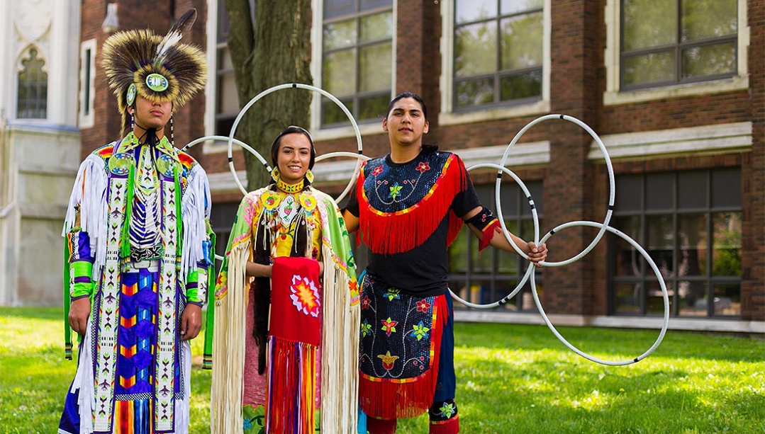 3 Indigenous dancers dressed in various regalia standing in front of Dillon Hall