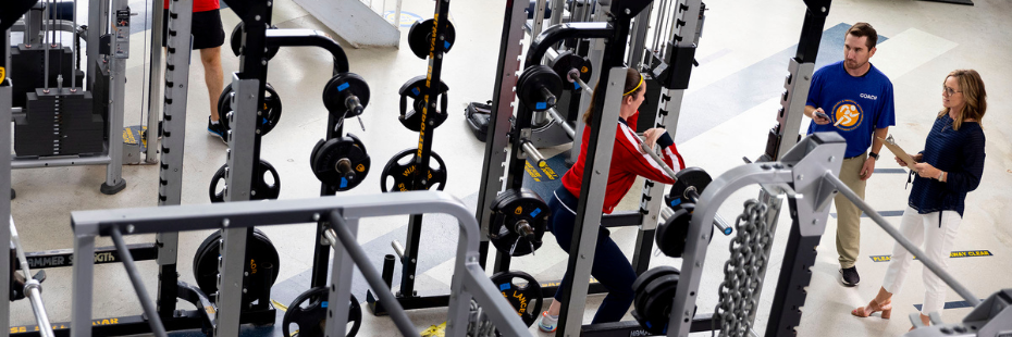 A male and female researcher look on as a female student works out in the gym.