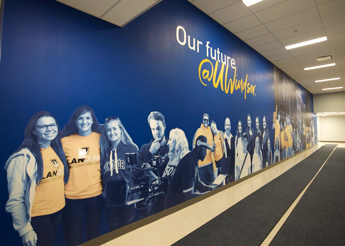 Blue wall with student photos in black, white and yellow read Our Future Made In Windsor. 