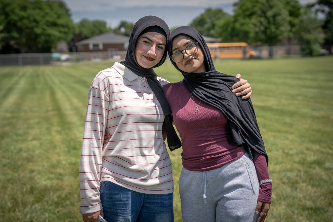Two high school girls in a hijabs, one wearing a white and red striped shirt and the other a burgundy shirt, smile for the camera
