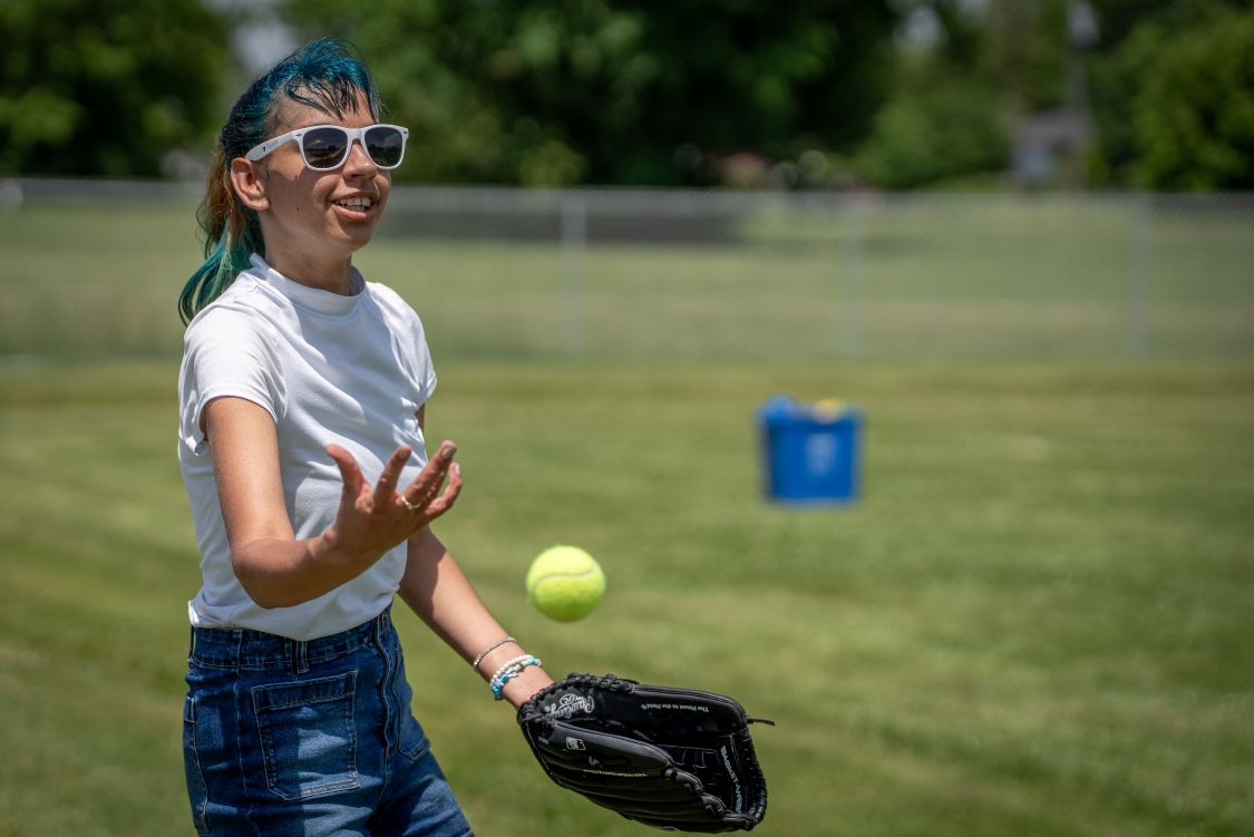 A teenage girl in sunglasses with blue hair catches a baseball