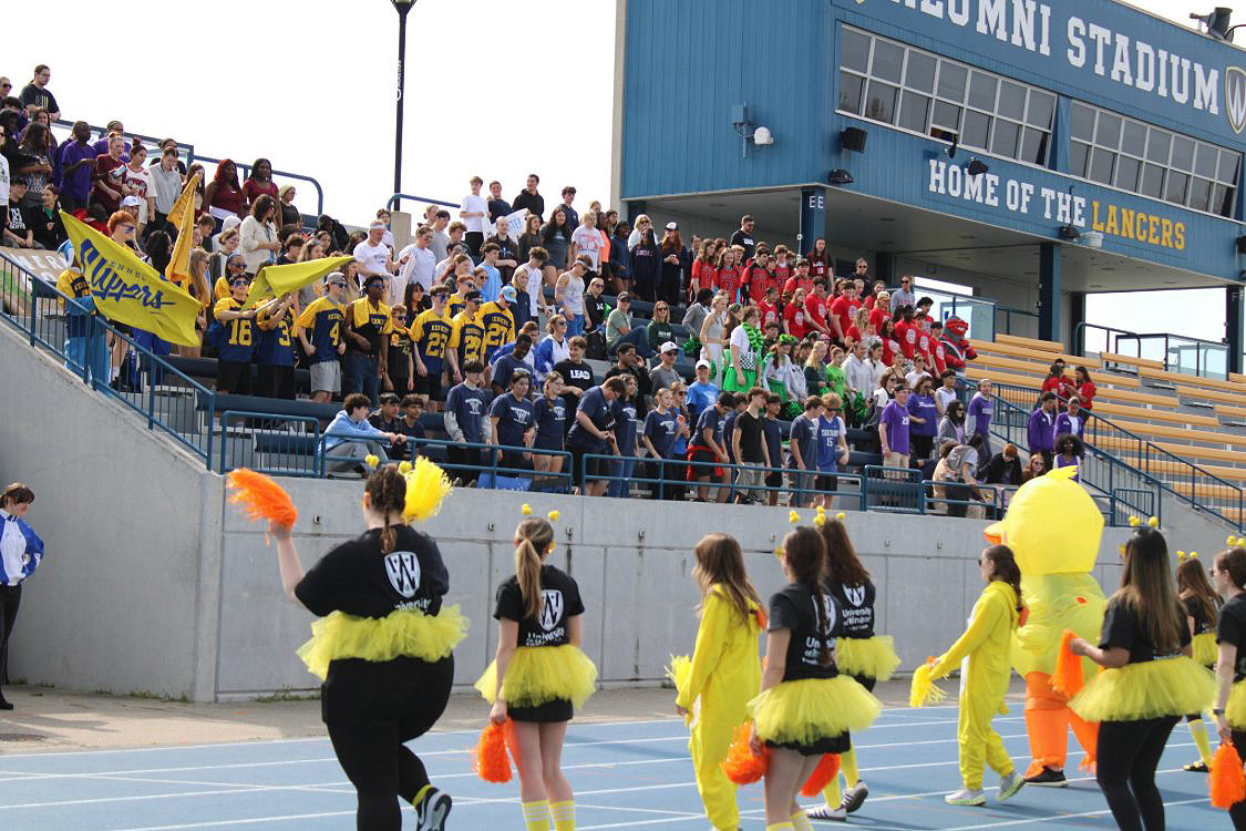 A group of teacher candidates and someone in a duck costume does a cheer performance in front of stands full of students at Alumni stadium