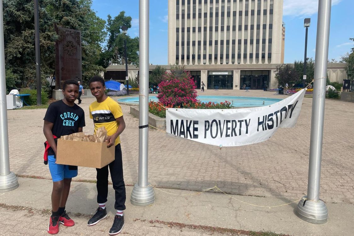 Two boys stand with a box of lunches in front of a sign that says Make Poverty History