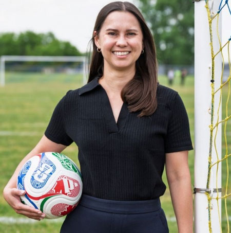 Kristen Morrison holds a soccer ball with soccer field behind her