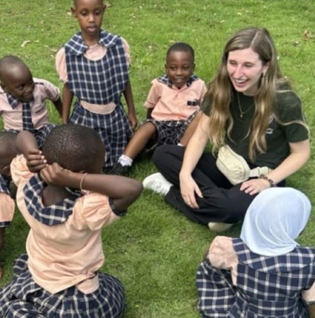 Smiling students and a UWindsor teacher candidate sitting in the grass in Tanzania