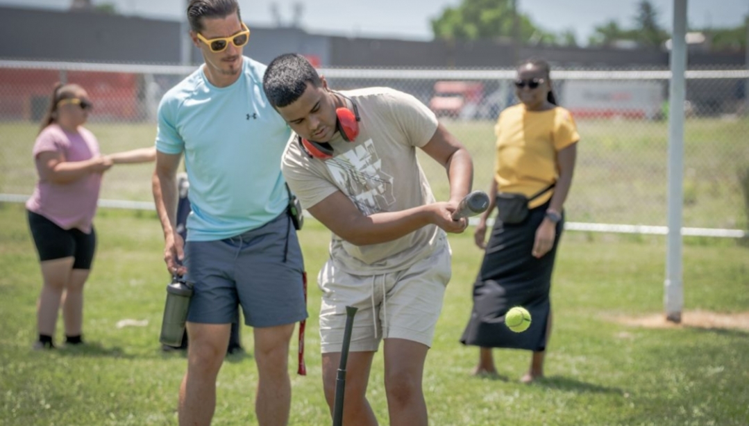 A teacher helps a student play baseball with other students in background