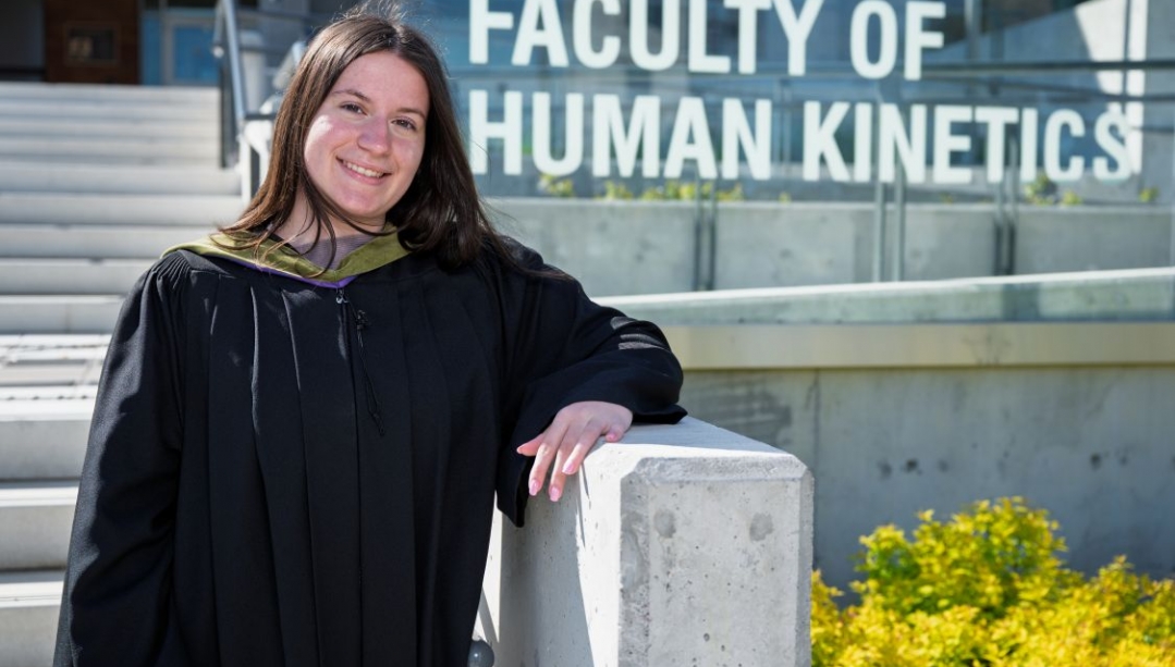 Sophia Boucas outside the sign for the Faculty of Human Kinetics wearing a graduation gown