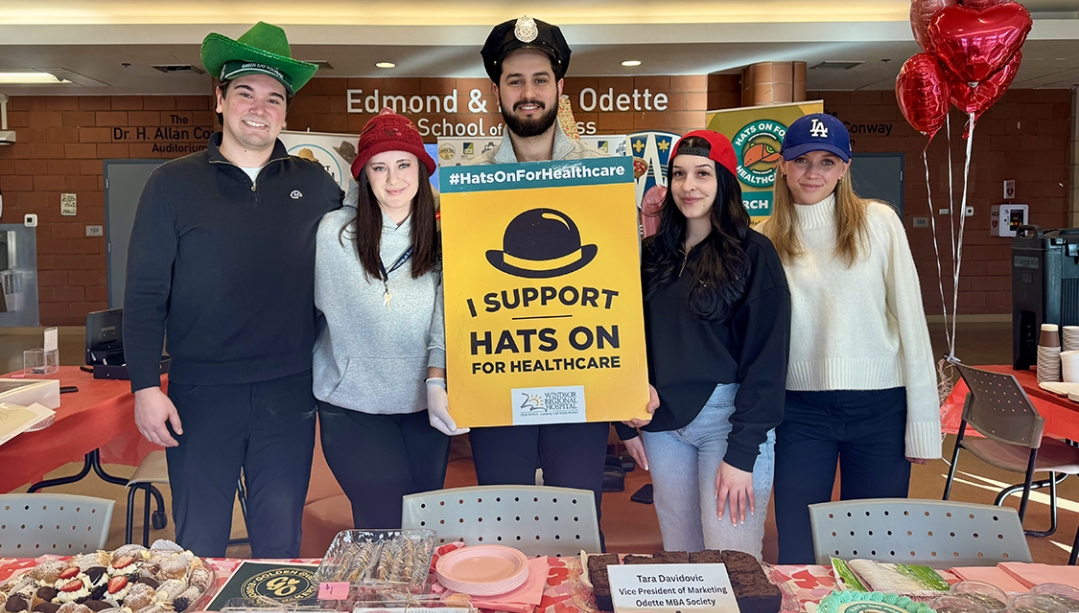 Odette School of Business MBA students hold up Hats on for Healthcare bake sale sign