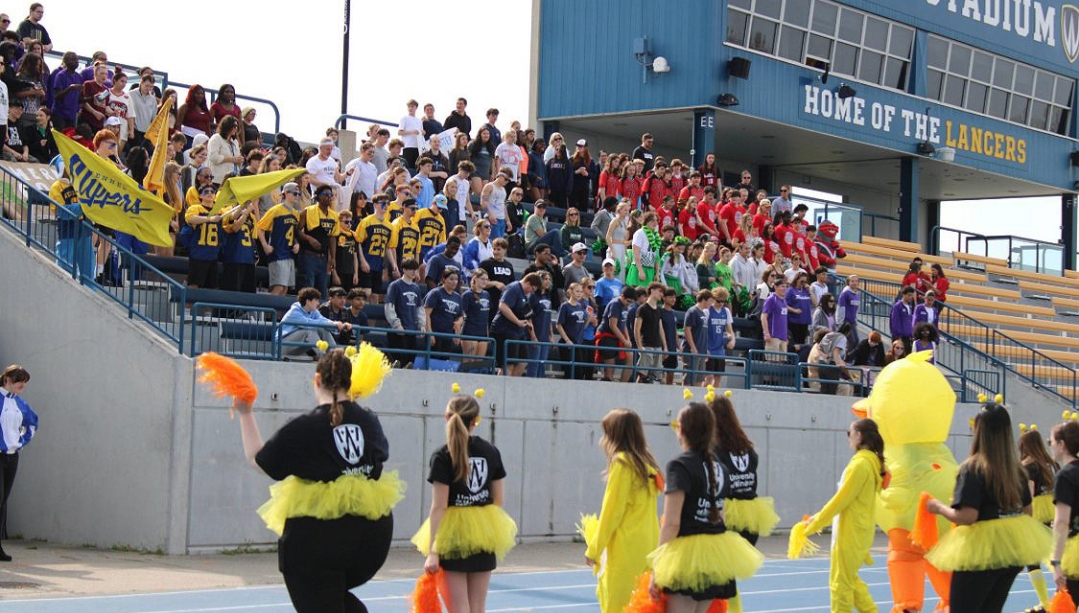A group of teacher candidates and someone in a duck costume does a cheer performance in front of stands full of students at Alumni stadium