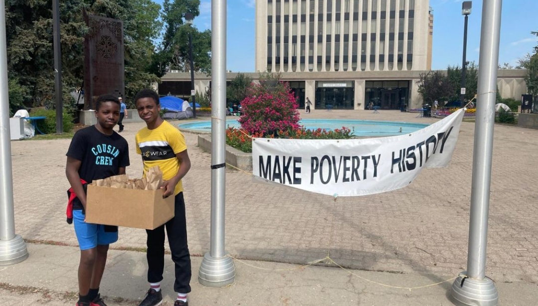 Two boys stand with a box of lunches in front of a sign that says Make Poverty History