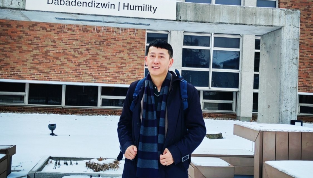 Marc Xu at UWindsor on Turtle Island Walk in front of signage with text Dabadendizwin Humility