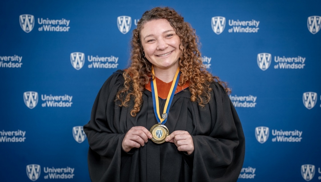 Kelsey Schumacher stands in front of a UWindsor backdrop in her grad gown holding her LEAD medal