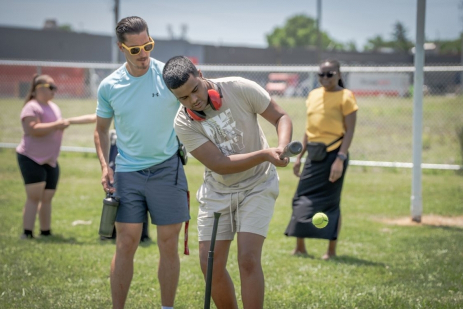 A teacher helps a student play baseball with other students in background