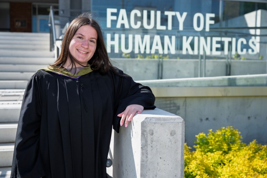 Sophia Boucas outside the sign for the Faculty of Human Kinetics wearing a graduation gown