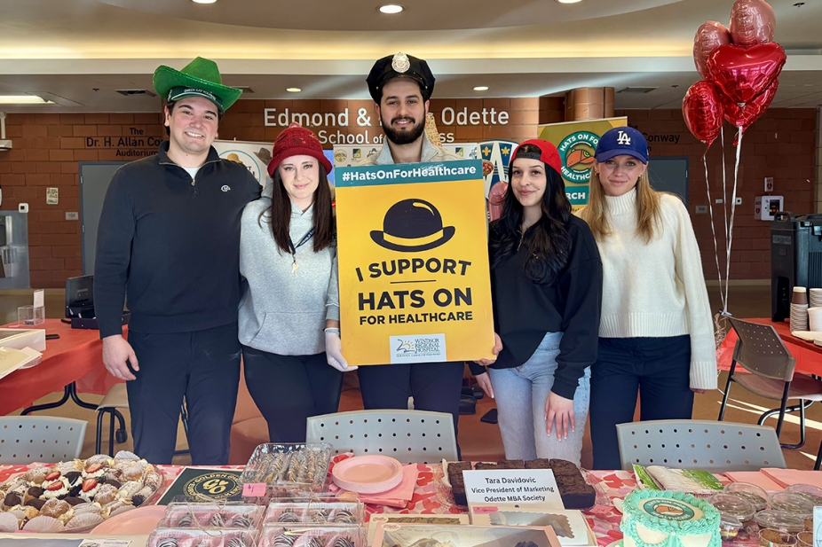 Odette School of Business MBA students hold up Hats on for Healthcare bake sale sign