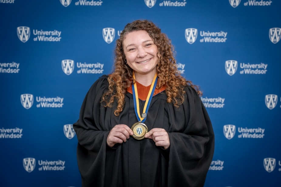 Kelsey Schumacher stands in front of a UWindsor backdrop in her grad gown holding her LEAD medal