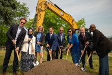 Eight people shovel dirt in front of a yellow excavator 