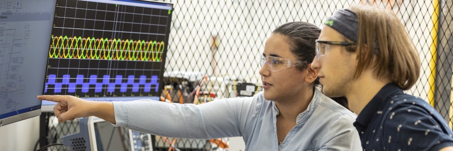 A man and a woman sitting in front of screens and discussing, the woman pointing at something on one of the screens
