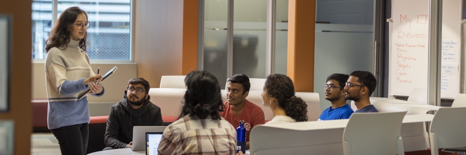 Student standing with a clipboard and speaking to a handful of people seated on couches around a table in a comfortable meeting room setting