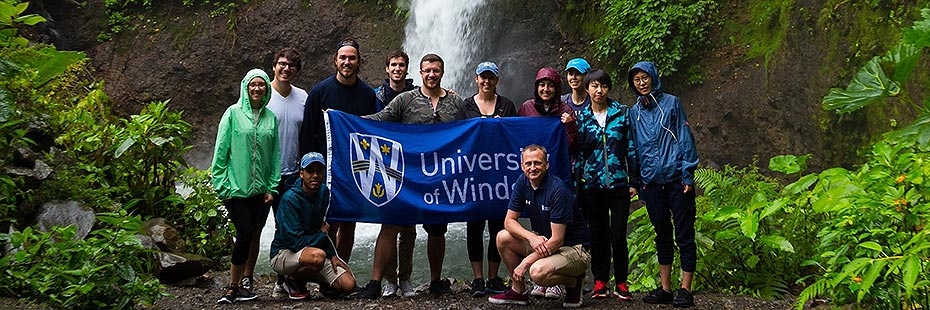 Instructors and students holding a UWindsor banner in front of Costa Rica waterfall