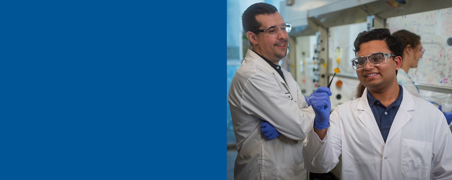 Two men in lab coats in a science lab look at a small square orange piece of paper