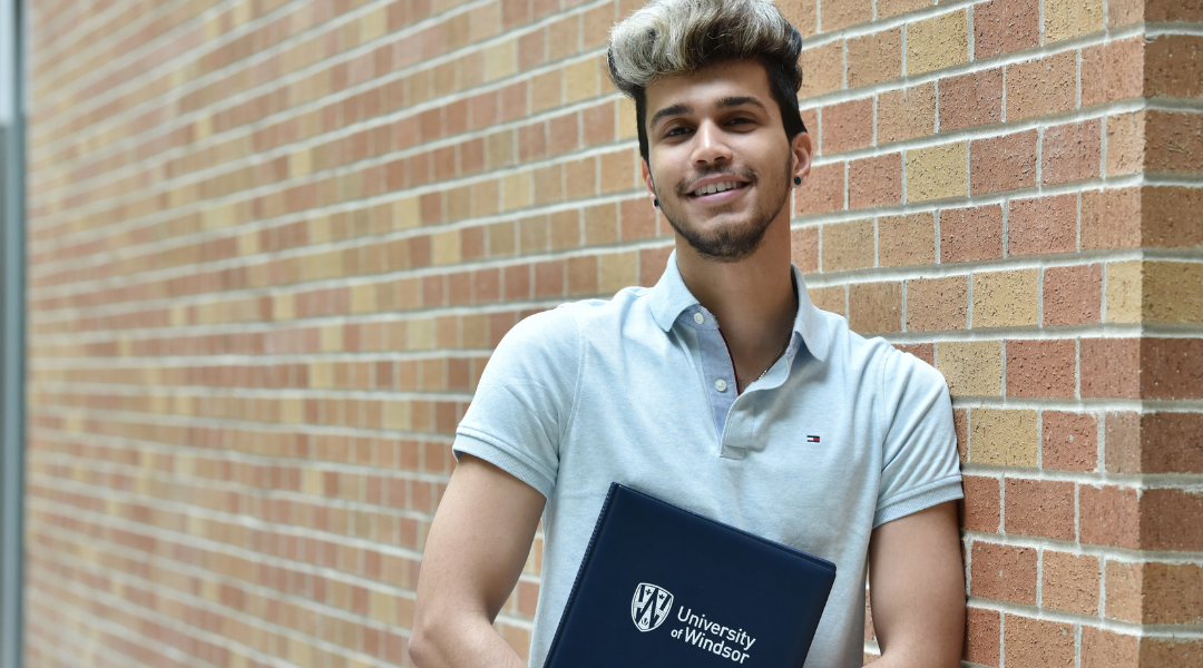 a student leaning against a wall holding a University of Windsor binder