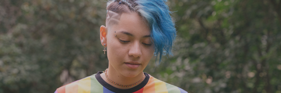 Young adult with blue hair wearing a rainbow t-shirt looking down from the camera